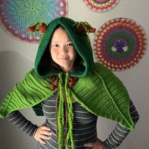 May include: A green crocheted hooded cape with brown and green fringe. The cape has a leaf-like design and two horns on the hood.