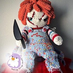Puede incluir: Muñeca de crochet con cabello rojo, ojos azules y una cara cosida. La muñeca lleva un mono azul y blanco con las palabras "Good Guys" en la parte delantera. La muñeca sostiene un cuchillo negro y está sentada sobre una superficie roja.