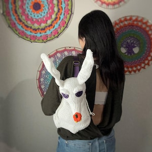 Può includere: Uno zaino bianco all'uncinetto a forma di cane con occhi viola e un naso arancione. Lo zaino è indossato da una persona con lunghi capelli neri. La persona è in piedi davanti a un muro con arazzi all'uncinetto colorati.