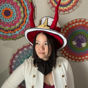 May include: A whimsical hat with a white crown, red horns, and a red brim. The hat is trimmed with white and black. The person is wearing a white jacket with gold buttons and a black bow tie. Colorful crochet wall hangings are in the background.