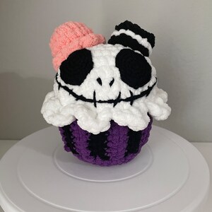 Peut inclure: Un objet en forme de cupcake crocheté avec un design Jack Skellington. Le cupcake a un dessus blanc avec un bord à volants, une base rayée violette et noire et un chapeau noir et blanc. Le visage a des yeux noirs et un sourire cousu.