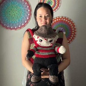 Pode incluir: Uma mulher está segurando um boneco de Freddy Krueger de crochê. O boneco está usando um chapéu marrom, uma camisa listrada vermelha e verde e calças pretas. O boneco tem um rosto branco com olhos pretos e uma boca vermelha.