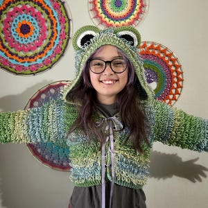 Puede incluir: Una chaqueta con capucha de ganchillo verde, azul y blanco con tem&aacute;tica de rana, con una capucha a juego con ojos y orejas grandes. La chaqueta tiene un cierre de cinta. En el fondo hay tapices de ganchillo coloridos.