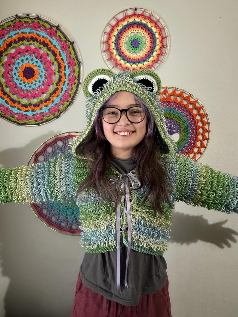 Puede incluir: Una chaqueta con capucha de ganchillo verde, azul y blanco con tem&aacute;tica de rana, con una capucha a juego con ojos y orejas grandes. La chaqueta tiene un cierre de cinta. En el fondo hay tapices de ganchillo coloridos.