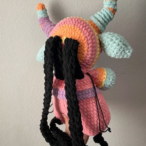 May include: A crocheted plush backpack in the shape of a monster with rainbow colored horns and a pink body. The backpack has black straps.