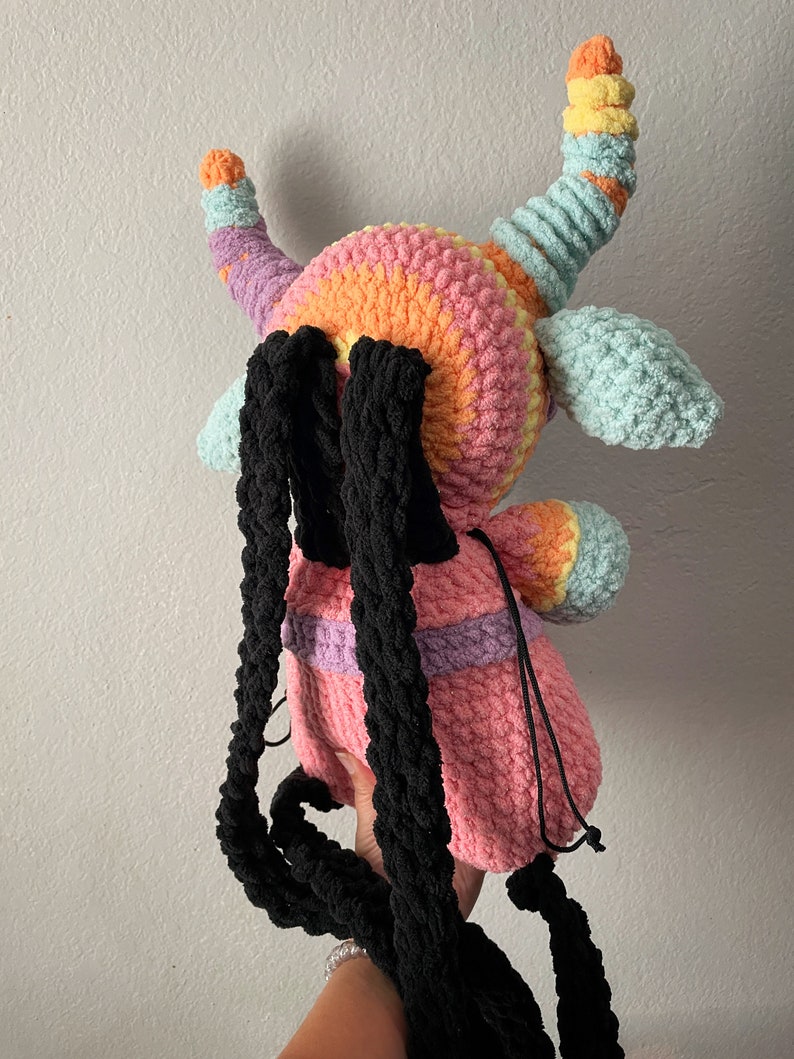 May include: A crocheted plush backpack in the shape of a monster with rainbow colored horns and a pink body. The backpack has black straps.