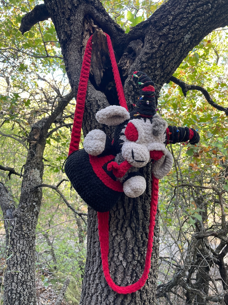 May include: A black and red crocheted bag with a gray and red crocheted plush toy attached to the strap. The bag and toy are hanging from a tree branch.