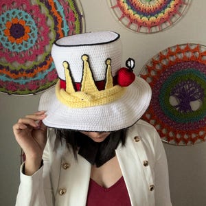 May include: A white crochet top hat with a black brim, a yellow crown, and a red apple accent. The hat is part of a costume. Decorative crochet wall hangings in various colors are in the background.