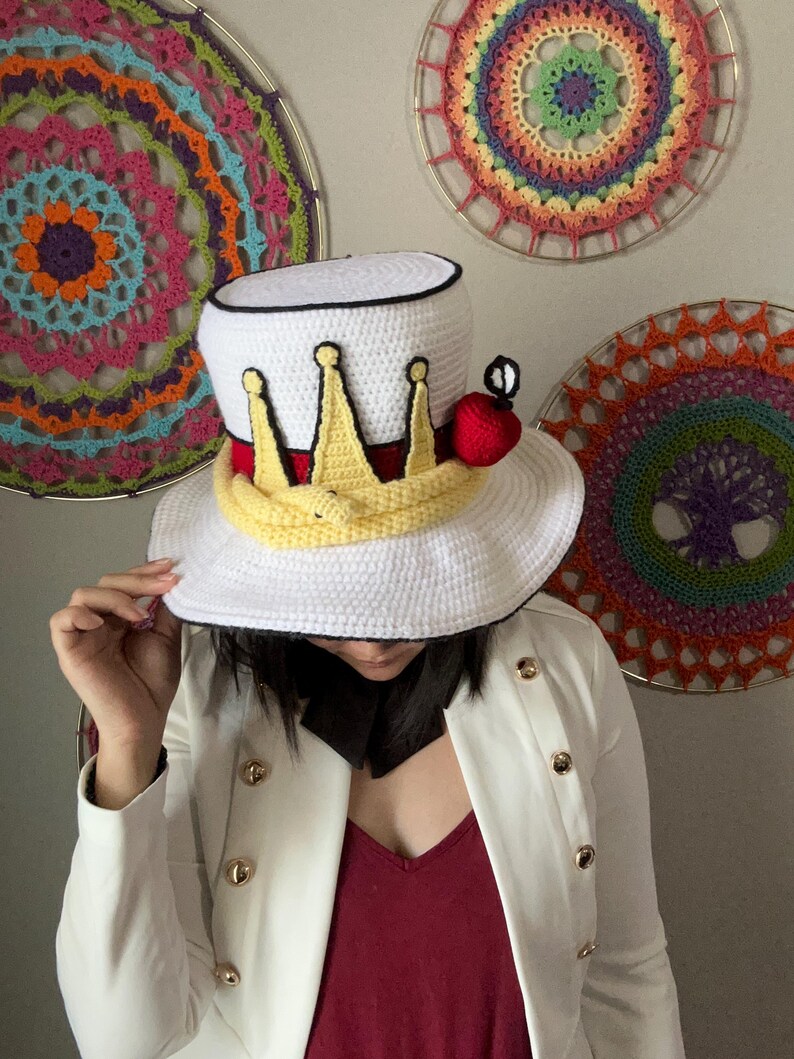 May include: A white crochet top hat with a black brim, a yellow crown, and a red apple accent. The hat is part of a costume. Decorative crochet wall hangings in various colors are in the background.