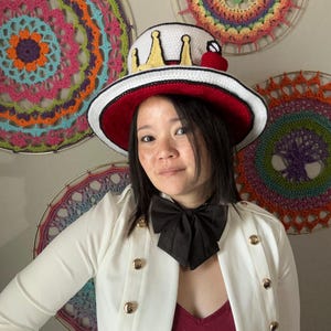 May include: A white and red brimmed hat with a crown design and a red cherry. The hat is worn with a white jacket with gold buttons and a black bow tie. Colorful crochet wall hangings are in the background.