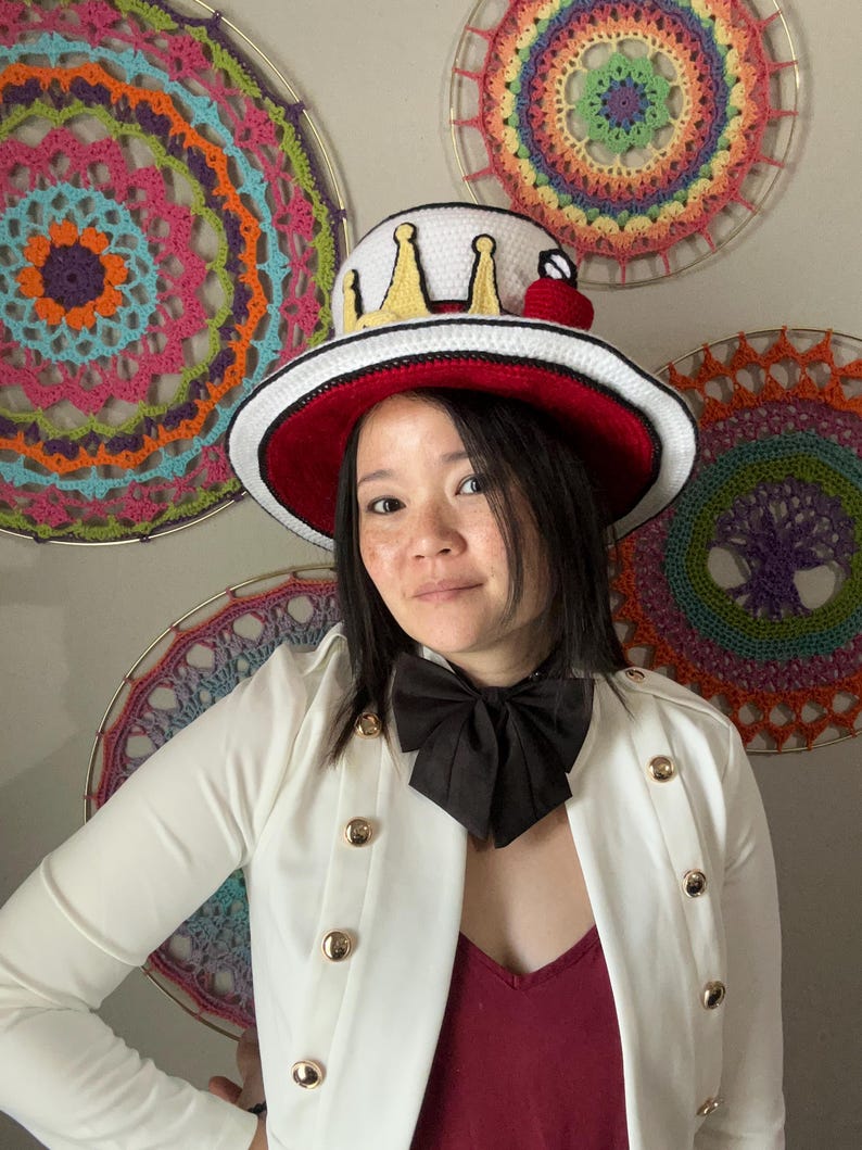May include: A white and red brimmed hat with a crown design and a red cherry. The hat is worn with a white jacket with gold buttons and a black bow tie. Colorful crochet wall hangings are in the background.