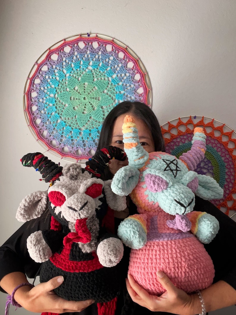 May include: Two crocheted plush toys in the shape of goats. One goat is pink and has a rainbow pattern, the other is gray and black with red accents. Both goats have horns and a pentagram on their foreheads.