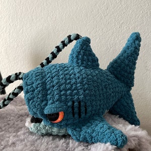 May include: A teal blue crocheted shark plush toy with black and white striped yarn for the tail. The shark has a large, red eye and a stitched-on mouth.