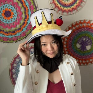 May include: A white top hat with a yellow crown design and a red apple accent. The hat has a wide brim and a red interior. The person is wearing a white jacket with gold buttons and a black necktie. Colorful crochet wall hangings are in the background.