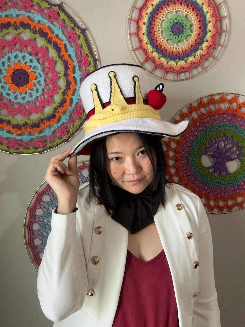 May include: A white top hat with a yellow crown design and a red apple accent. The hat has a wide brim and a red interior. The person is wearing a white jacket with gold buttons and a black necktie. Colorful crochet wall hangings are in the background.