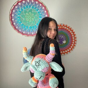 May include: A crocheted backpack shaped like a goat with rainbow colored yarn. The backpack has a pentagram on its forehead and a pink, purple, and orange color scheme. The backpack is being worn by a person with long black hair.