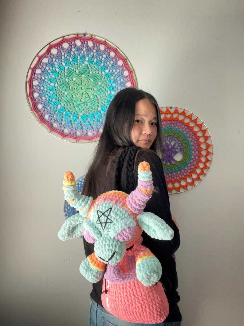 May include: A crocheted backpack shaped like a goat with rainbow colored yarn. The backpack has a pentagram on its forehead and a pink, purple, and orange color scheme. The backpack is being worn by a person with long black hair.