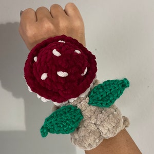 May include: A handmade crochet mushroom wristband. The mushroom cap is burgundy with white spots, the gills are white, and the leaves are green. The wristband is beige.