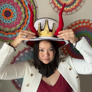 May include: A white hat with a red brim, adorned with a gold crown and red horns. The hat is being held up. The person is wearing a white blazer with gold buttons and a black bow tie. Colorful crochet art is in the background.