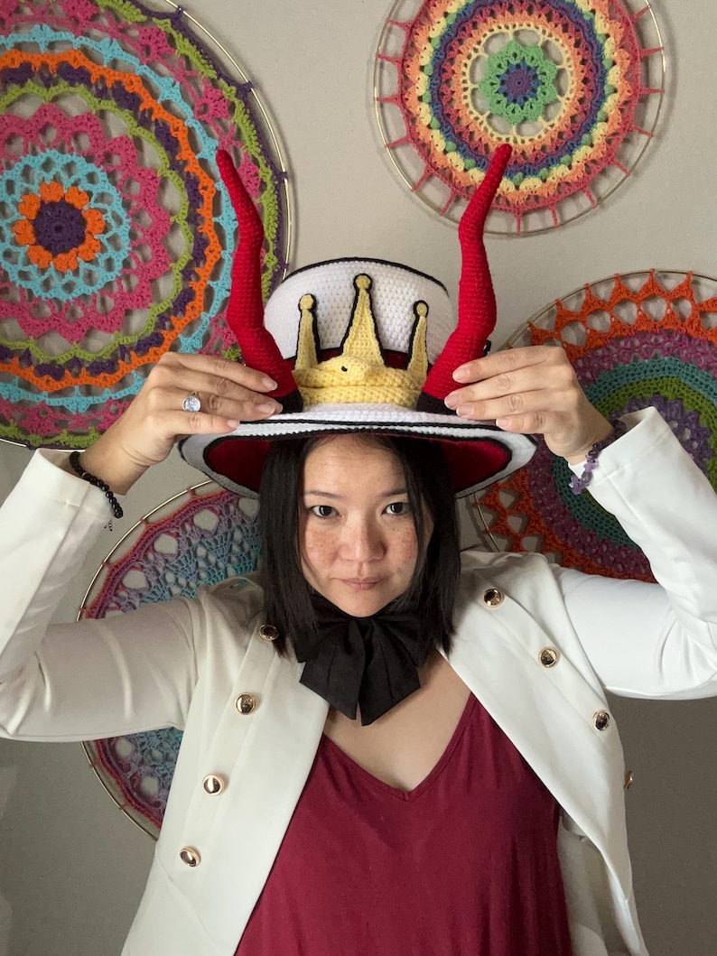 May include: A white hat with a red brim, adorned with a gold crown and red horns. The hat is being held up. The person is wearing a white blazer with gold buttons and a black bow tie. Colorful crochet art is in the background.
