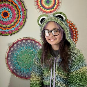 Puede incluir: Una sudadera con capucha de rana de ganchillo verde y azul con capucha y orejas a juego. La chaqueta tiene un corte corto y un cierre de cinta. Tres mandalas de ganchillo coloridos cuelgan de la pared detr&aacute;s de la chaqueta.