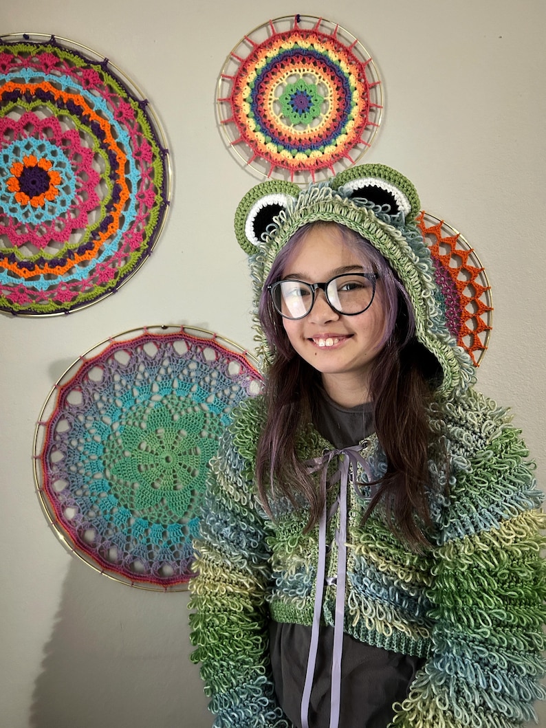 Puede incluir: Una sudadera con capucha de rana de ganchillo verde y azul con capucha y orejas a juego. La chaqueta tiene un corte corto y un cierre de cinta. Tres mandalas de ganchillo coloridos cuelgan de la pared detr&aacute;s de la chaqueta.
