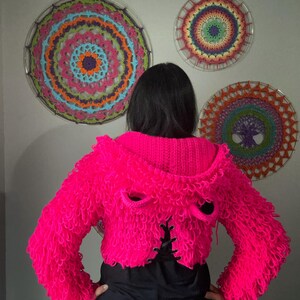 Puede incluir: Una chaqueta corta rosa brillante y difusa con capucha y detalles de ojales. La chaqueta se usa sobre una camiseta negra. Tres coloridos tapices de pared circulares con dise&ntilde;os intrincados est&aacute;n en el fondo.