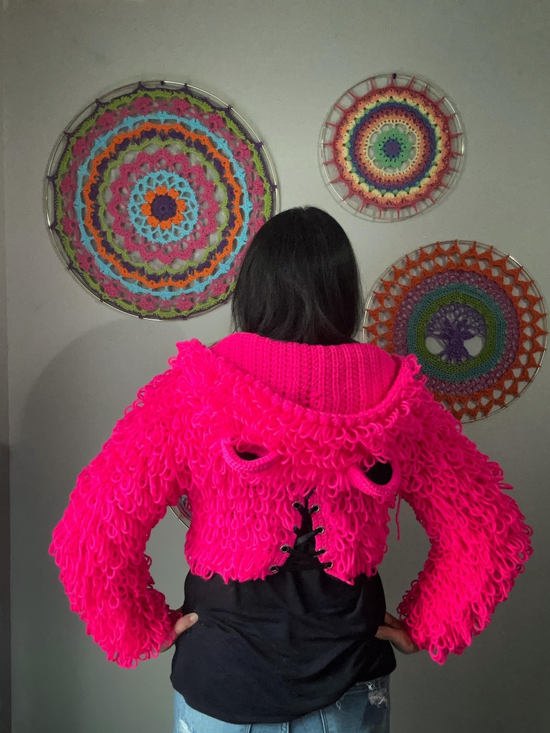 Puede incluir: Una chaqueta corta rosa brillante y difusa con capucha y detalles de ojales. La chaqueta se usa sobre una camiseta negra. Tres coloridos tapices de pared circulares con dise&ntilde;os intrincados est&aacute;n en el fondo.