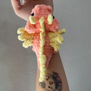 May include: A pink and yellow crocheted dragon toy with black eyes. The dragon is sitting on a person's arm.
