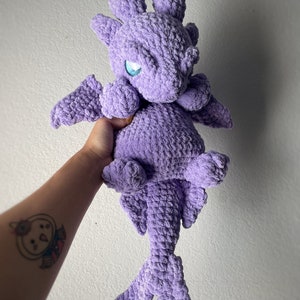 Può includere: Un giocattolo di peluche di drago viola all'uncinetto con occhi blu. Il drago ha una coda lunga e ali.