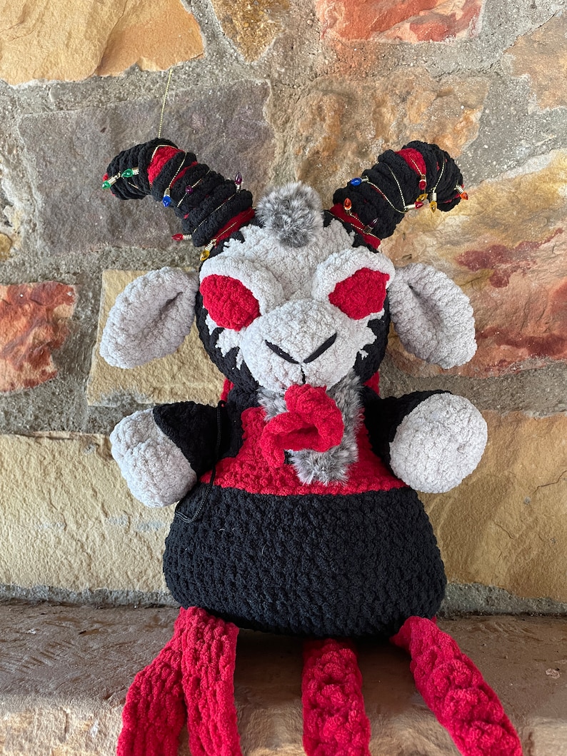 May include: A plush toy goat with red eyes, black and red fur, and red yarn horns. The goat is sitting on a stone surface.