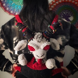 May include: A person wearing a black and white patterned sweater with a crocheted plush backpack in the shape of a white creature with red eyes and a red mouth. The backpack has black and red crocheted details.