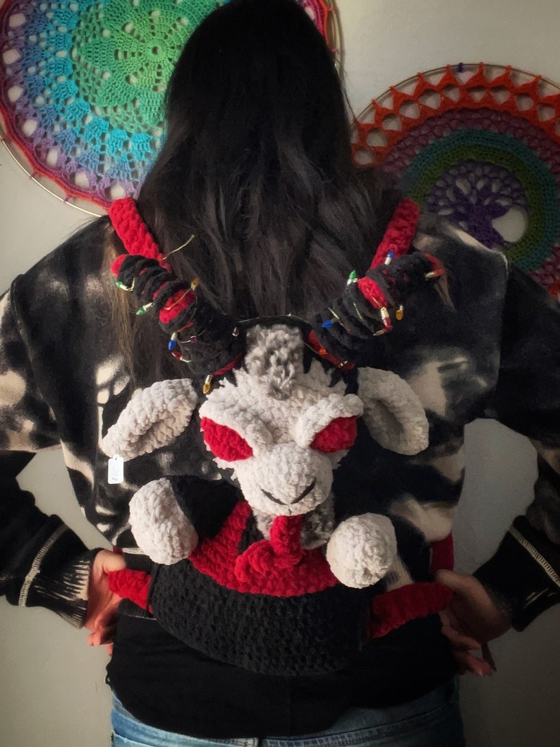 May include: A person wearing a black and white patterned sweater with a crocheted plush backpack in the shape of a white creature with red eyes and a red mouth. The backpack has black and red crocheted details.