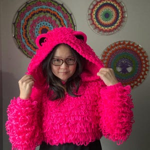 Puede incluir: Una sudadera con capucha corta rosa brillante hecha a mano con orejas de oso. La sudadera con capucha tiene un dise&ntilde;o texturizado en bucle y se usa con una falda negra. Decoraciones de pared circulares y coloridas en el fondo.