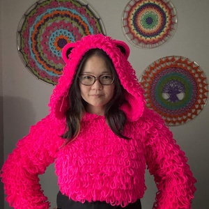Puede incluir: Una sudadera con capucha rosa brillante y difusa con orejas de oso. El su&eacute;ter corto tiene mangas largas y un dise&ntilde;o texturizado en bucle. La capucha est&aacute; levantada y la persona lleva gafas. Tres coloridos tapices circulares est&aacute;n en el fondo.