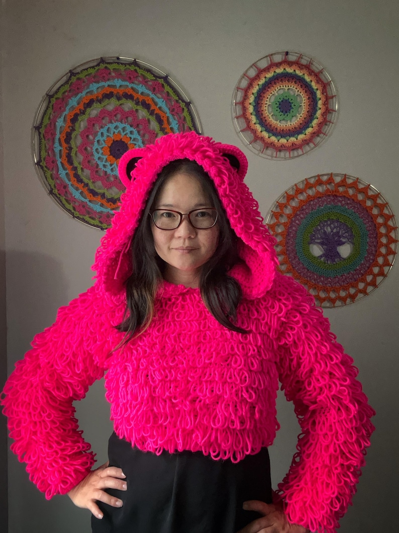 Puede incluir: Una sudadera con capucha rosa brillante y difusa con orejas de oso. El su&eacute;ter corto tiene mangas largas y un dise&ntilde;o texturizado en bucle. La capucha est&aacute; levantada y la persona lleva gafas. Tres coloridos tapices circulares est&aacute;n en el fondo.