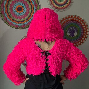 Puede incluir: Una chaqueta corta con capucha de color rosa vibrante con un dise&ntilde;o texturizado en bucle. La chaqueta presenta un cuello en V con cordones de cinta negra. En el fondo, hay colgantes de pared circulares decorativos y coloridos.