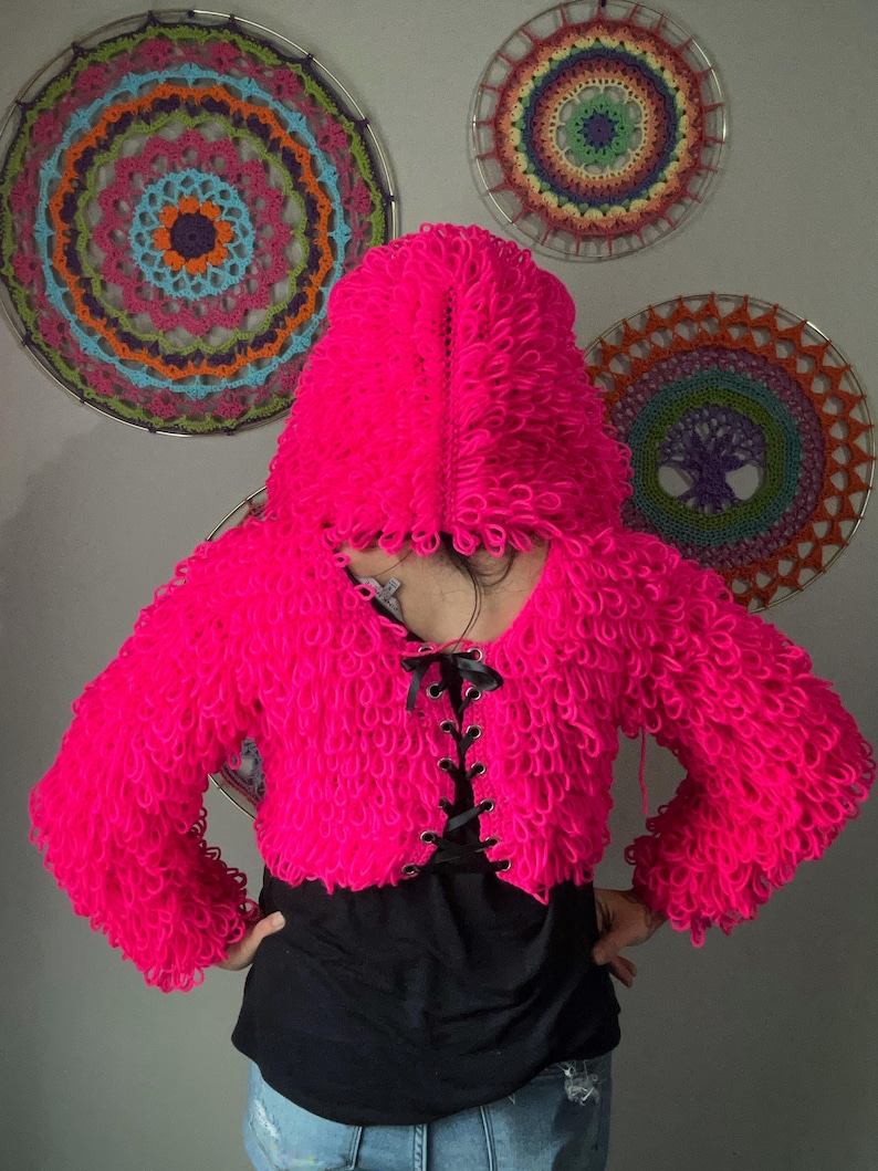 Puede incluir: Una chaqueta corta con capucha de color rosa vibrante con un dise&ntilde;o texturizado en bucle. La chaqueta presenta un cuello en V con cordones de cinta negra. En el fondo, hay colgantes de pared circulares decorativos y coloridos.
