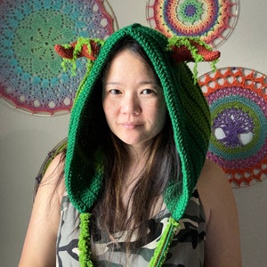 Puede incluir: Una persona lleva una capucha de dragón de crochet verde con largas borlas verdes. La capucha tiene cuernos rojos y hojas verdes. La persona lleva un top de tirantes de camuflaje.