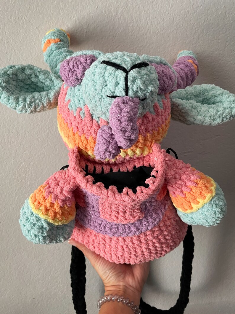 May include: A crocheted stuffed animal shaped like a monster with a pink, purple, orange, and light blue color scheme. The monster has a black embroidered face and a black drawstring bag.