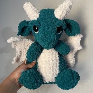 May include: A crocheted plush dragon toy with teal and white fur. The dragon has large black eyes and white horns.