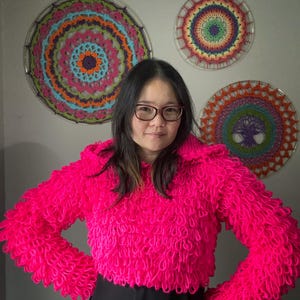 Puede incluir: Un su&eacute;ter corto rosa brillante y texturizado con mangas largas. El su&eacute;ter tiene un dise&ntilde;o de bucle. Tres tapices de pared circulares y coloridos est&aacute;n en el fondo. Los tapices de pared tienen un dise&ntilde;o de ganchillo con varios colores.