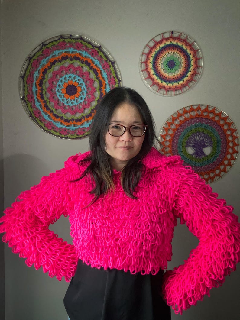 Puede incluir: Un su&eacute;ter corto rosa brillante y texturizado con mangas largas. El su&eacute;ter tiene un dise&ntilde;o de bucle. Tres tapices de pared circulares y coloridos est&aacute;n en el fondo. Los tapices de pared tienen un dise&ntilde;o de ganchillo con varios colores.