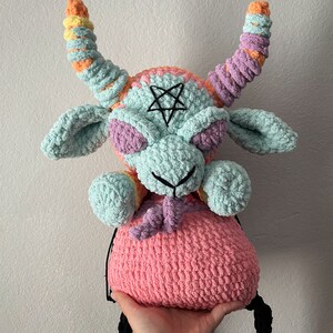 May include: A crocheted plush toy shaped like a goat with a pink, purple, and blue color scheme. The goat has a pentagram on its forehead and long, colorful horns. The toy is holding a pink crocheted bag.
