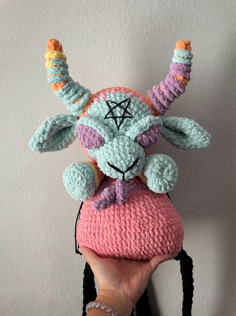 May include: A crocheted plush toy shaped like a goat with a pink, purple, and blue color scheme. The goat has a pentagram on its forehead and long, colorful horns. The toy is holding a pink crocheted bag.