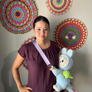 Może przedstawiać: Kobieta nosi torbę na ramię w kolorze jasnofioletowym. Torba zawiera niebieską pluszową zabawkę z kapturem królika i zielonymi skrzydłami. Na ścianie jako dekoracja wiszą szydełkowe mandale w różnych kolorach.