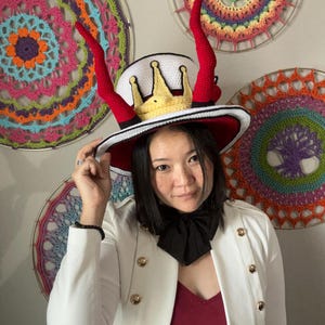 May include: A whimsical, handmade hat with a white crown, a gold crown, and red horns. The hat has a red brim and a black and white band. The hat is a unique accessory, perfect for costume parties or themed events.