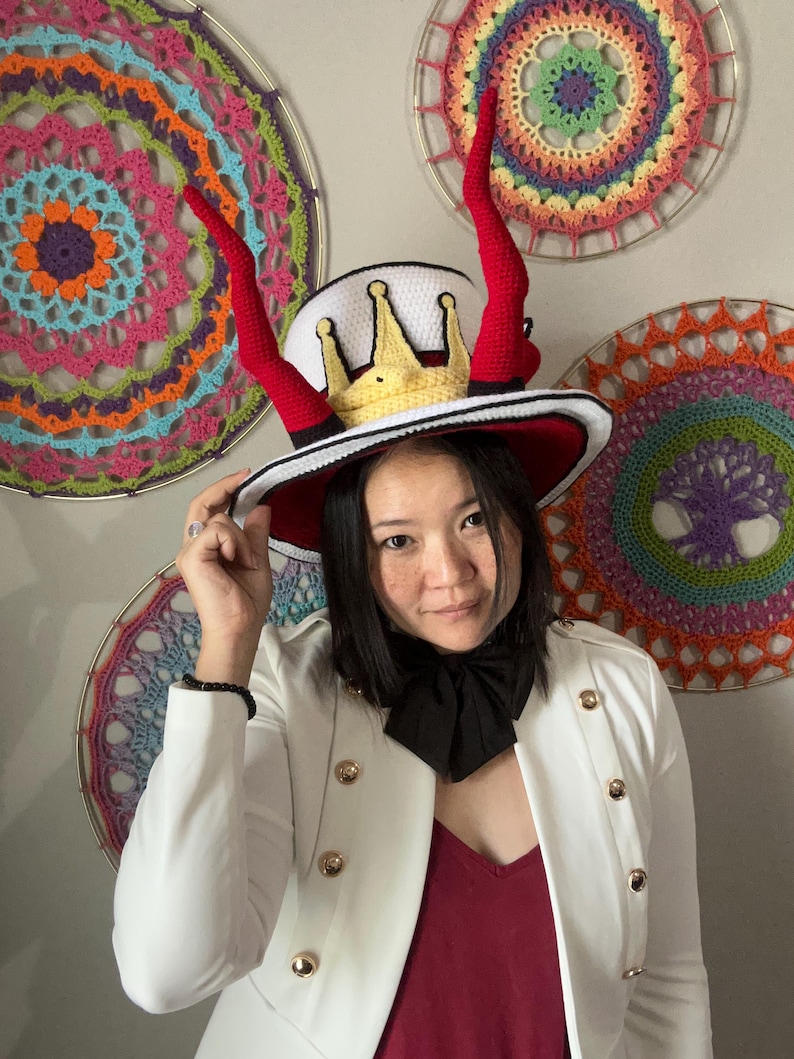 May include: A whimsical, handmade hat with a white crown, a gold crown, and red horns. The hat has a red brim and a black and white band. The hat is a unique accessory, perfect for costume parties or themed events.