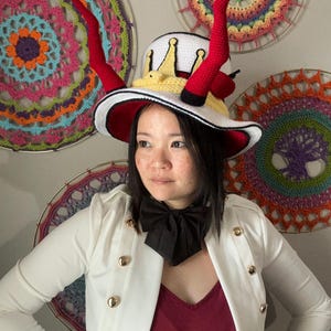 May include: A whimsical white hat with a red interior and brim, adorned with red horns and a yellow crown. The hat is trimmed with black and white accents. The person is wearing a white jacket with gold buttons and a black bow tie.