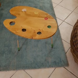 Puede incluir: Una mesa de madera en forma de paleta de artista con un acabado de madera clara. La mesa tiene cuatro patas y un agujero redondo en el centro de la paleta. La paleta tiene cinco salpicaduras de pintura de diferentes colores: blanco, amarillo, rojo, verde y negro.